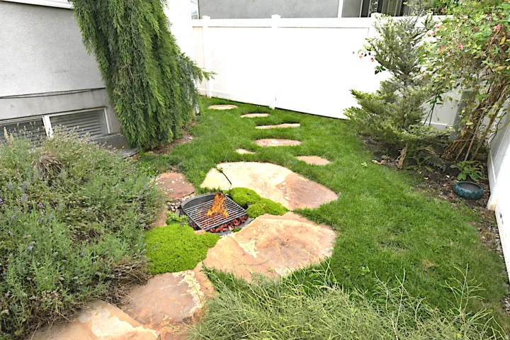 Landscaped side yard featuring a fire pit, stepping stone path, mature greenery, and a white privacy fence