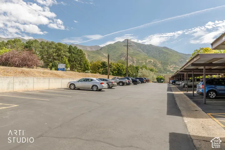 Paved parking lot with covered parking structures and a retaining wall, bordered by mature trees and a mountain backdrop