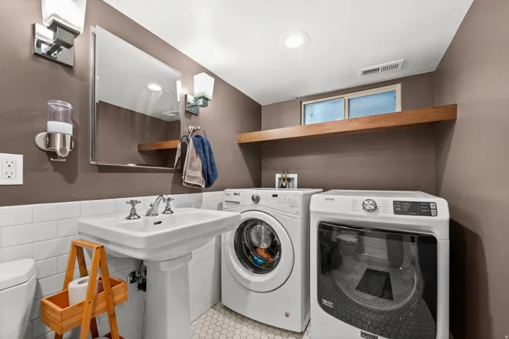 Pedestal sink with chrome fixtures, subway tile wainscoting, wall-mounted lighting, hexagon tile flooring, and integrated laundry appliances