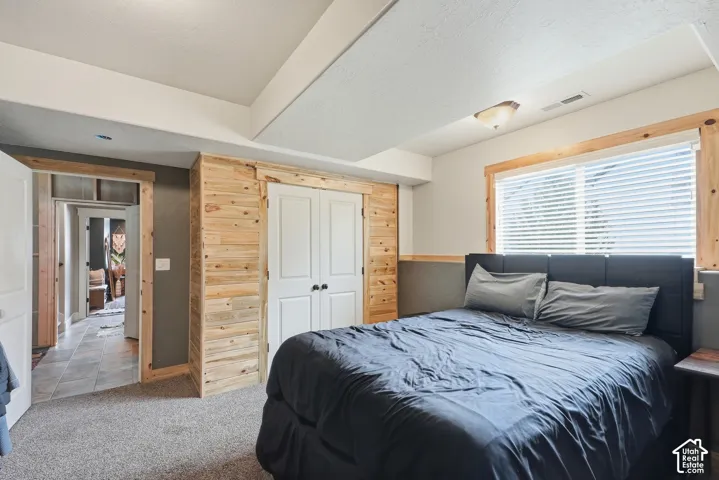 Carpeted room featuring a large window with blinds, wood-paneled accent wall, and double-door closet