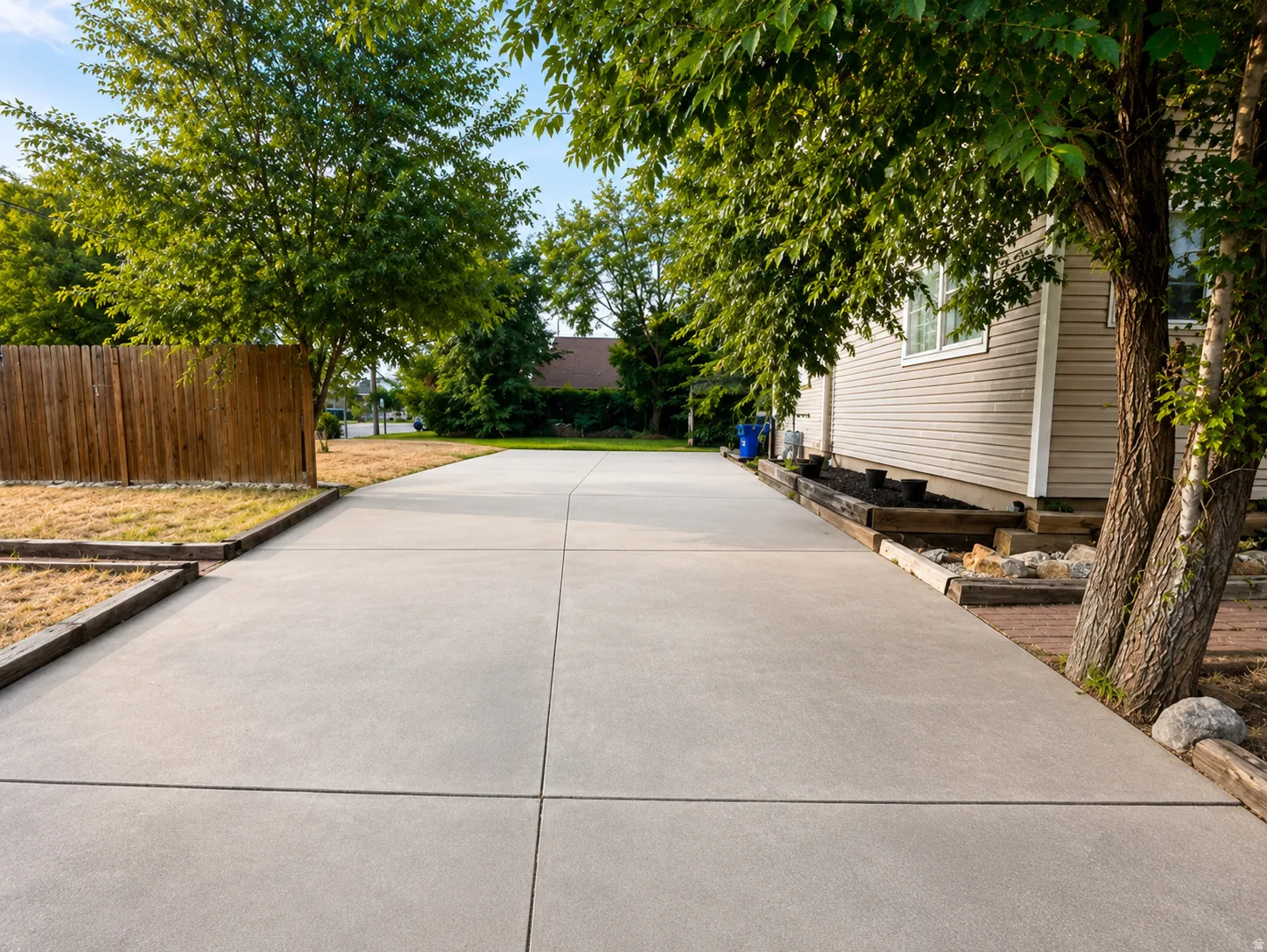 Extended concrete driveway with a wood privacy fence, mature trees, and a raised garden bed with timber framing