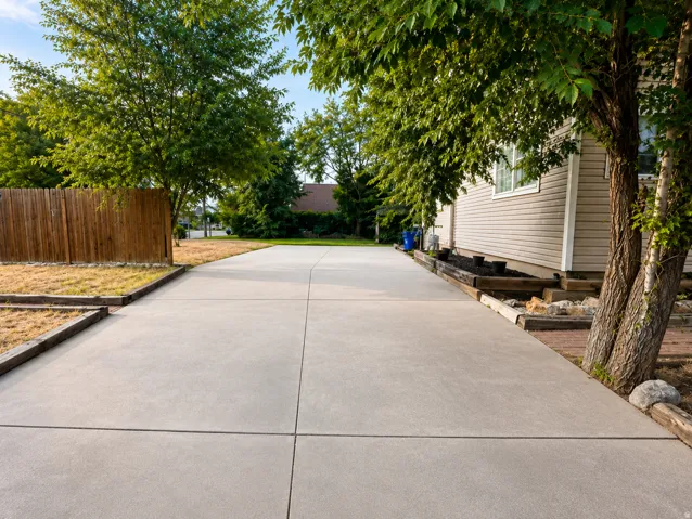 Extended concrete driveway with a wood privacy fence, mature trees, and a raised garden bed with timber framing