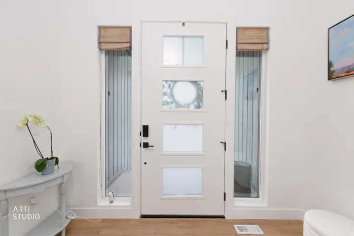 White entry door with frosted glass panels and matte black hardware, flanked by two full-length sidelights with Roman shades, set against white walls and wood-finish flooring
