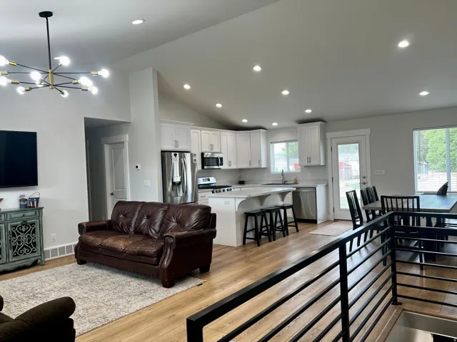 Open-concept living area featuring wood-finish flooring, vaulted ceilings, recessed lighting, and a modern kitchen with white cabinetry and stainless steel appliances