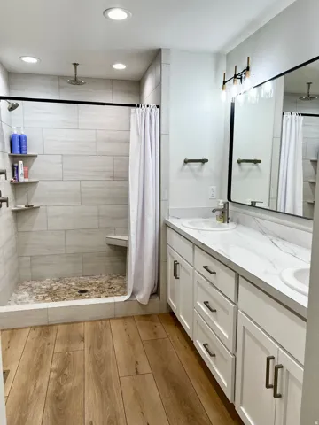 Spacious bathroom featuring wood-finish flooring, a dual-sink vanity with white cabinetry and light-toned countertops, and a walk-in shower with tiled walls and a pebble floor