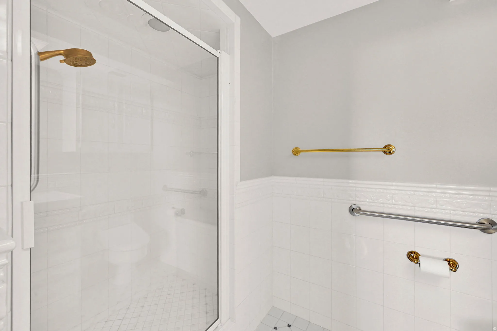 Glass-enclosed shower with a gold-tone shower head, white tiled walls, and mosaic tile flooring