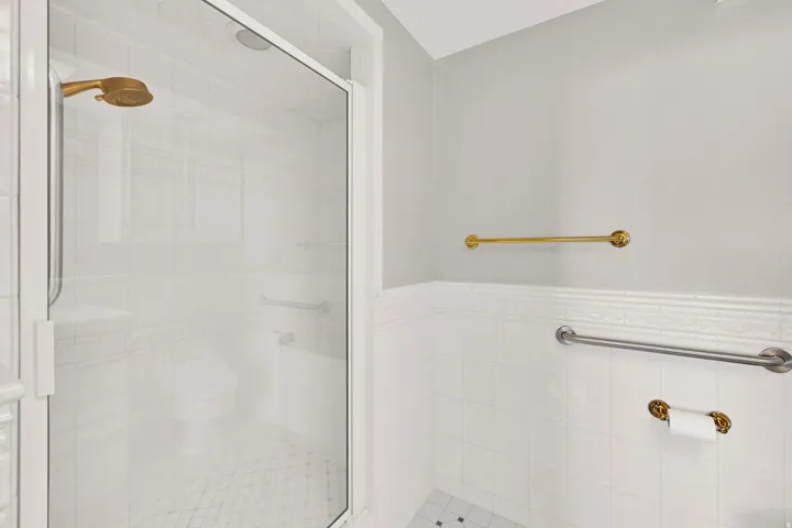 Glass-enclosed shower with a gold-tone shower head, white tiled walls, and mosaic tile flooring