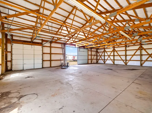 Spacious concrete-floor interior featuring exposed wood beam construction, multiple overhead fluorescent light fixtures, and a large roll-up garage door