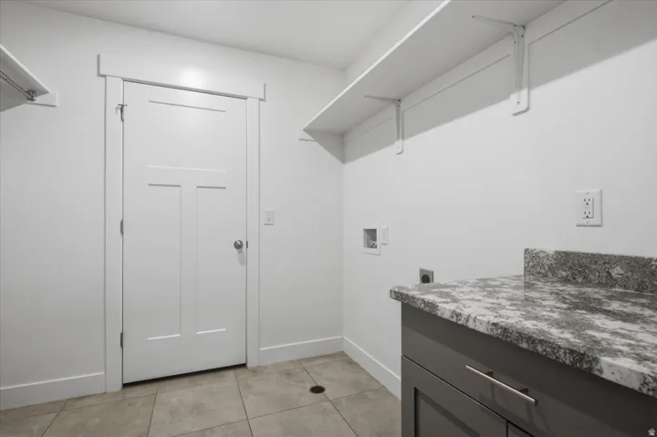 Laundry room featuring tile flooring, white shelving, gray cabinetry with metal hardware, and a stone countertop