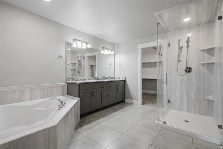 Dual-sink vanity with stone countertop, large soaking tub, walk-in shower with glass enclosure, tiled flooring, and recessed lighting