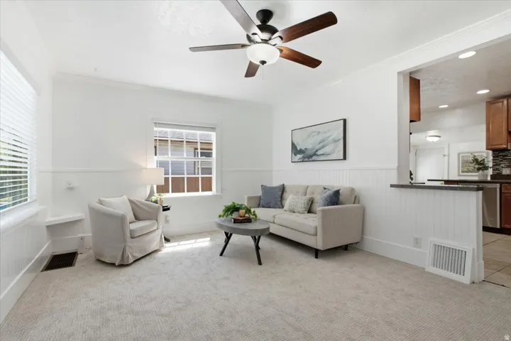 Bright living space with light-toned carpet, white wainscoting, and a ceiling fan for added comfort and character.