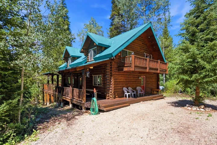 Log cabin exterior with a teal metal roof, a full-length covered porch, an upper-level balcony, and a gravel driveway