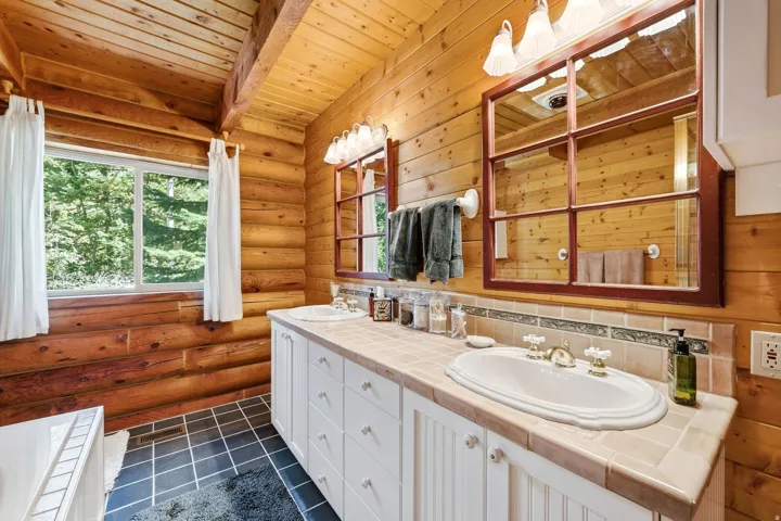 Wood-paneled bathroom featuring a dual vanity with white cabinetry, tile countertop, and a window