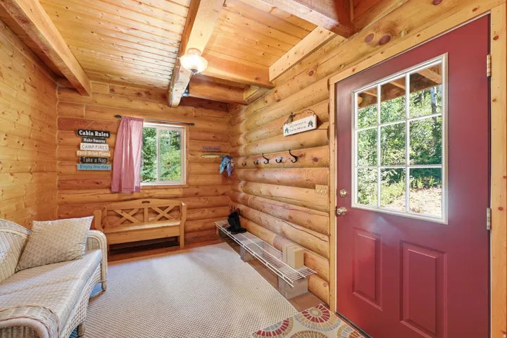 Log wall construction with exposed ceiling beams, a red paneled door with divided lites, a built-in wooden bench, and a small window with outdoor scenery