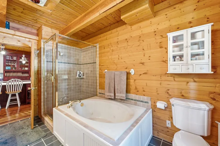 Wood-paneled walls and ceiling, corner soaking tub with tile surround, glass-enclosed shower with tiled walls, and a wall-mounted storage cabinet