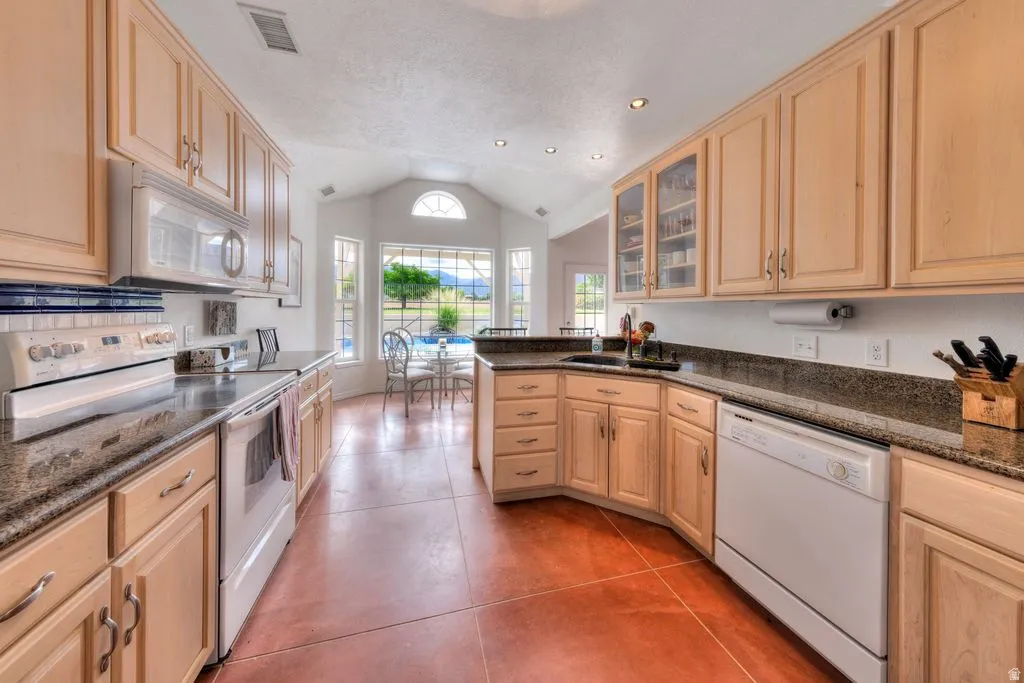 Galley kitchen featuring natural wood cabinetry with glass-front upper storage, stainless steel appliances, granite countertops, recessed lighting, and terracotta tile flooring