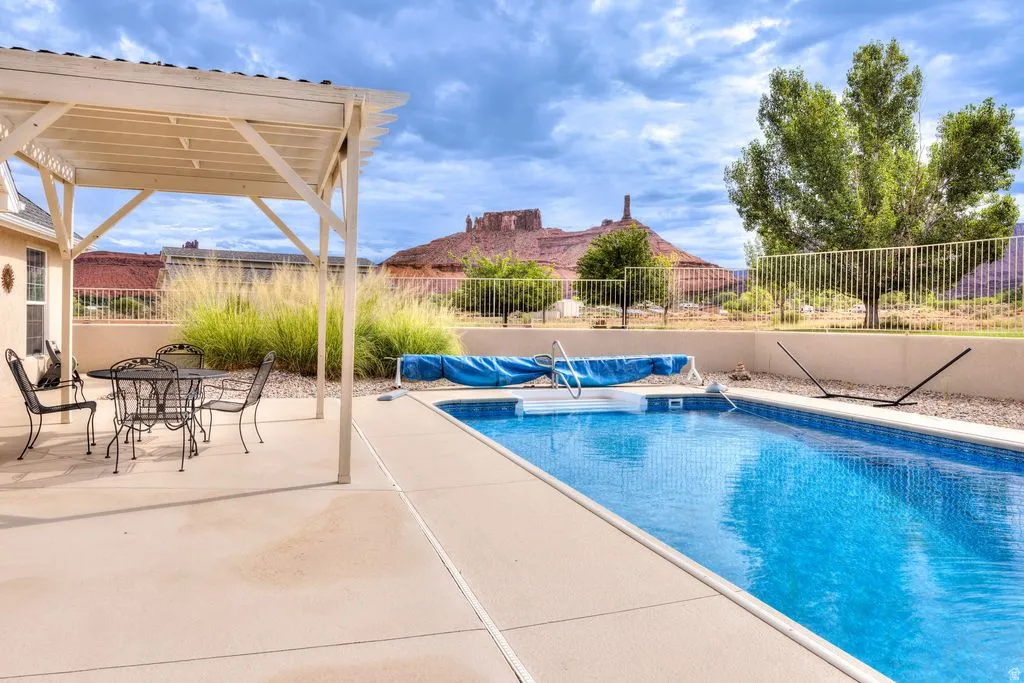 Outdoor swimming pool with a surrounding concrete patio, covered pergola, and a visible distant mountain formation
