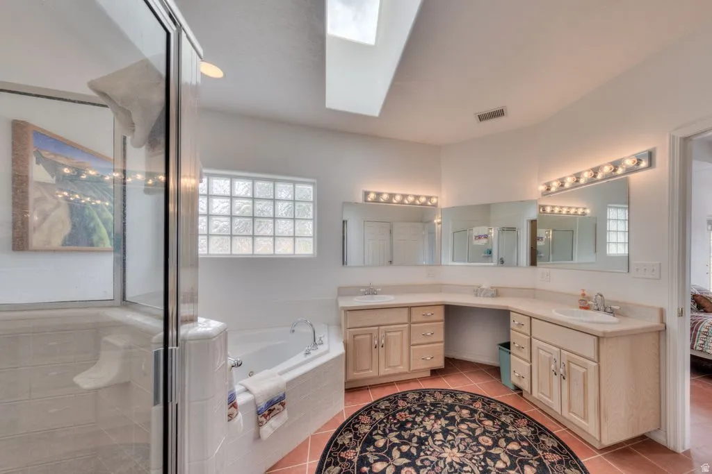 Dual vanity sinks with light wood-finish cabinetry, a jetted corner tub, a glass-enclosed shower, and a skylight