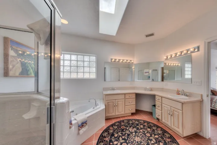 Dual vanity sinks with light wood-finish cabinetry, a jetted corner tub, a glass-enclosed shower, and a skylight