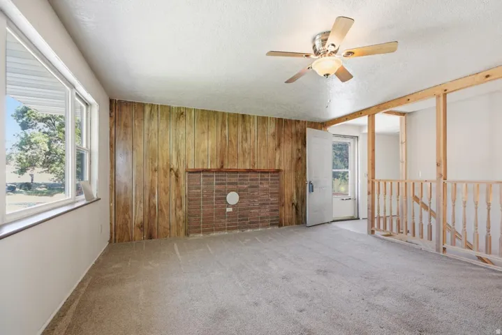 Wood panel accent wall with brick hearth, multiple windows, ceiling fan with light fixture, and carpeted flooring