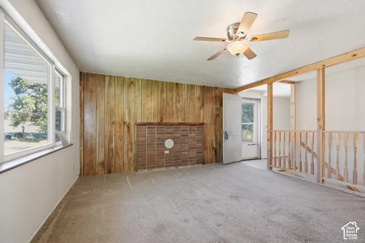 Wood panel accent wall with brick hearth, multiple windows, ceiling fan with light fixture, and carpeted flooring