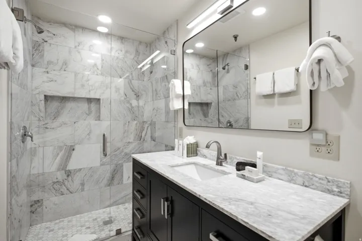 Spa-inspired bathroom featuring marble-look tile surround with integrated shower niche, polished stone countertop with undermount sink, matte black hardware, large illuminated mirror, and dark cabinetry with ample storage beneath the vanity