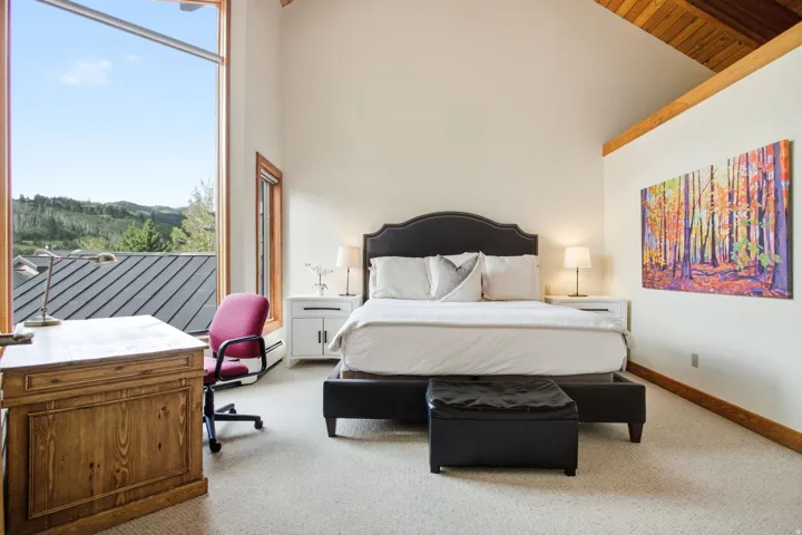 Vaulted ceiling with exposed wood beams, expansive window framing mountain scenery, wood-framed windows, built-in wooden desk, and light-colored carpeted flooring