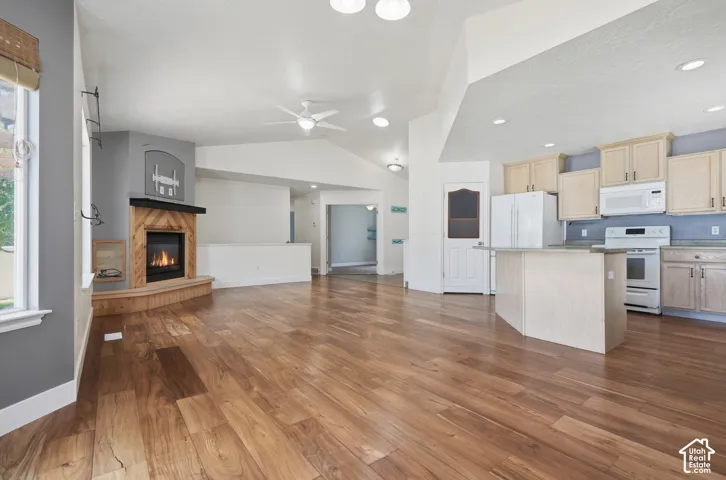 Expansive wood-finish flooring throughout, vaulted ceiling, built-in fireplace with a wood-clad surround, kitchen island with countertop, and white kitchen appliances