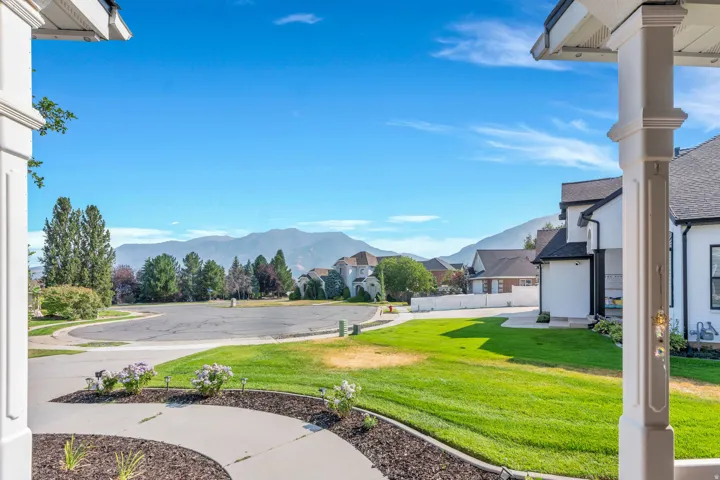 Expansive front yard with manicured lawn and mature trees, mountain backdrop, paved sidewalk with landscaping, and a broad street
