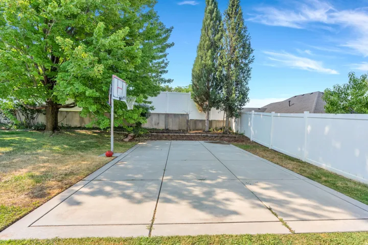 Paved concrete court with a basketball hoop, surrounded by a white privacy fence and mature trees