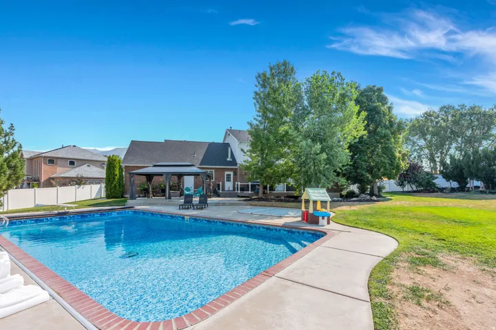 Large backyard featuring an in-ground swimming pool with brick coping, expansive concrete decking, a covered gazebo, and mature trees