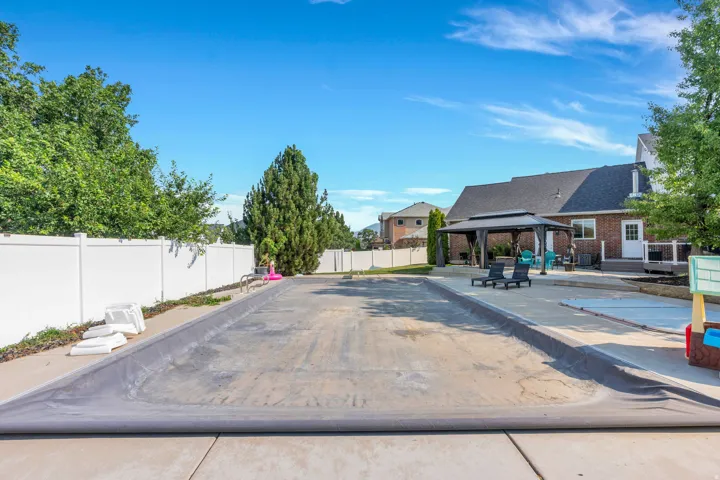 Covered in-ground pool, expansive concrete patio, outdoor gazebo, white vinyl fencing, and mature landscaping