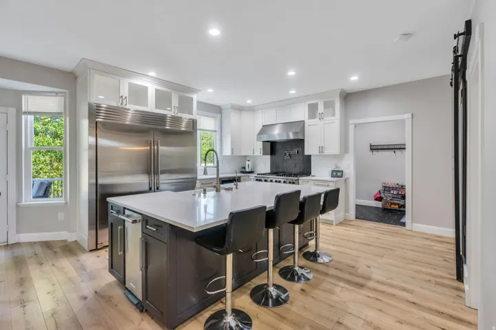 Modern kitchen featuring wood-finish flooring, stainless steel appliances, a large center island with seating, white upper cabinetry, and a dark tile backsplash