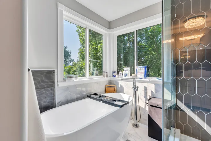 Freestanding soaking tub with floor-mounted faucet, corner windows, large format hexagon wall tile, walk-in shower with glass enclosure, and recessed lighting