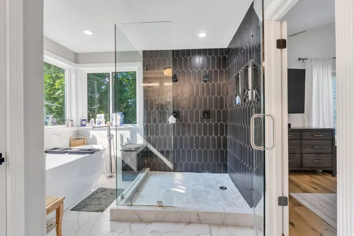 Oversized soaking tub with a bay window, glass-enclosed shower with dark picket tile, dual built-in shower niches, and recessed lighting