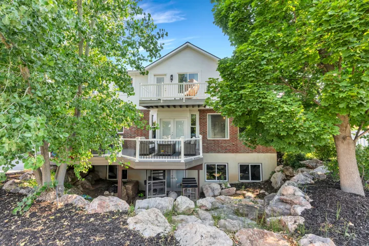 Multi-level exterior with white siding and brick accents, featuring two elevated balconies with white railings, and a rock-lined terraced landscape with mature trees