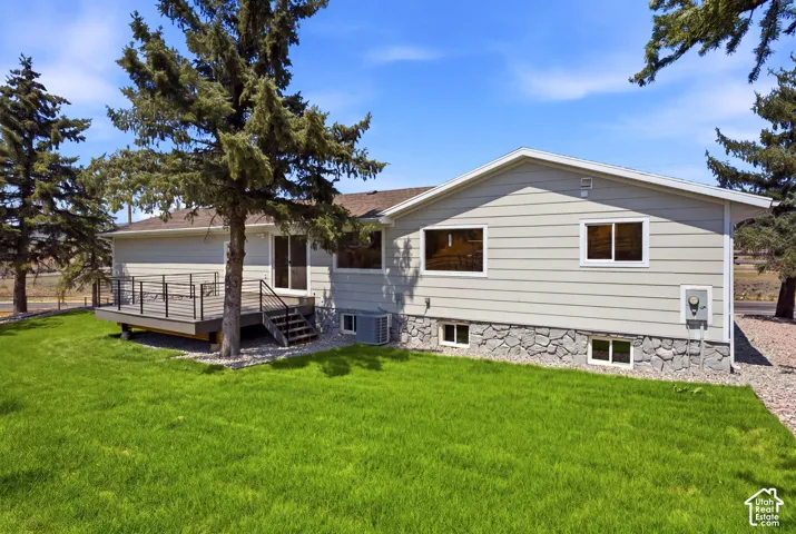 Expansive green lawn, multi-level deck with black metal railing, sliding glass door access, stone foundation, and light-toned siding
