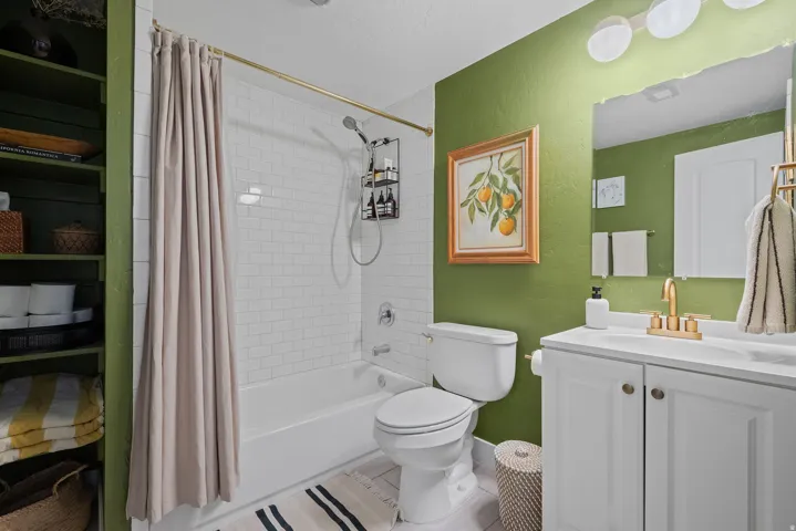White vanity with gold-tone faucet, white subway tile shower surround, built-in shelving, white bathtub, and toilet
