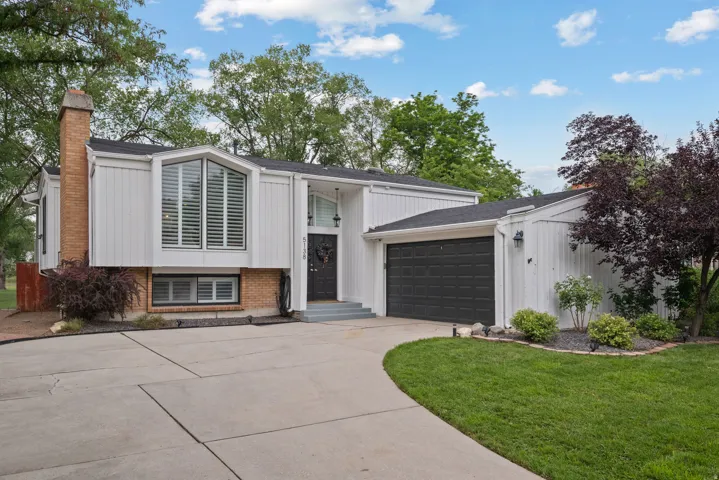 White siding exterior with a prominent brick chimney, large driveway, two-car garage, and mature landscaping