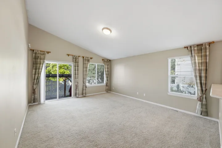 Vaulted ceiling, neutral wall paint, light-toned carpeting, sliding glass patio door, and multiple windows