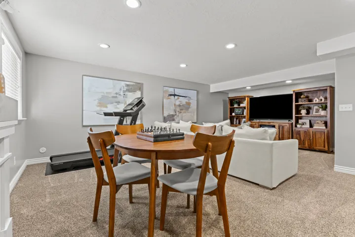 Versatile recreation room featuring a round wood-finish dining table with seating for four, recessed ceiling lighting, neutral carpet flooring, and built-in cabinetry with shelving and media storage