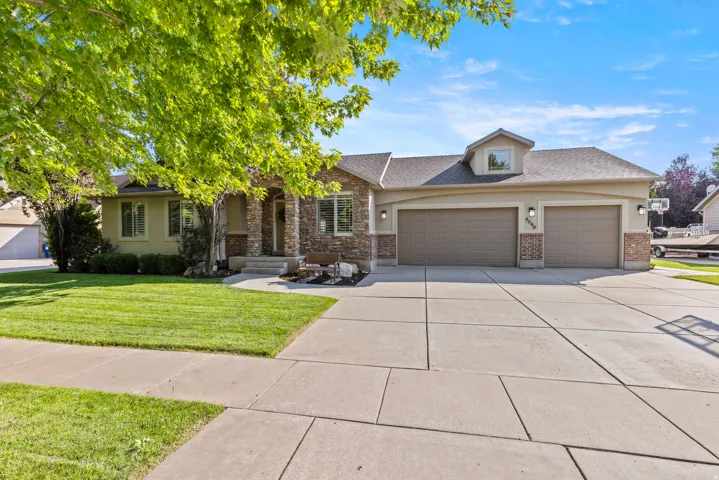 Welcoming front elevation featuring a stucco and stone exterior with a three-car garage, dormer window detail, and mature landscaping with established shade trees