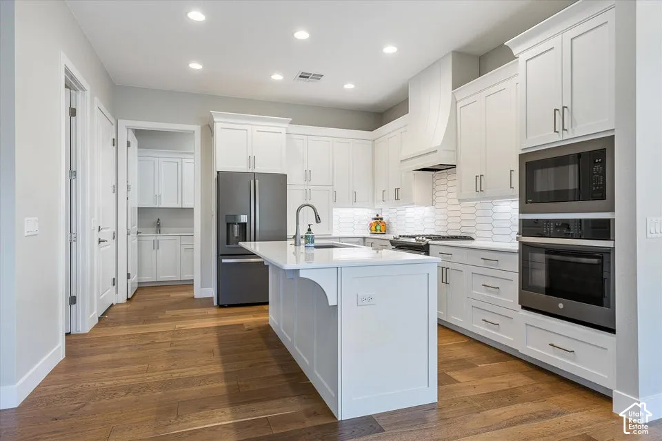 White shaker cabinetry, wood-finish flooring, recessed lighting, built-in stainless steel appliances, and a central island with a sink