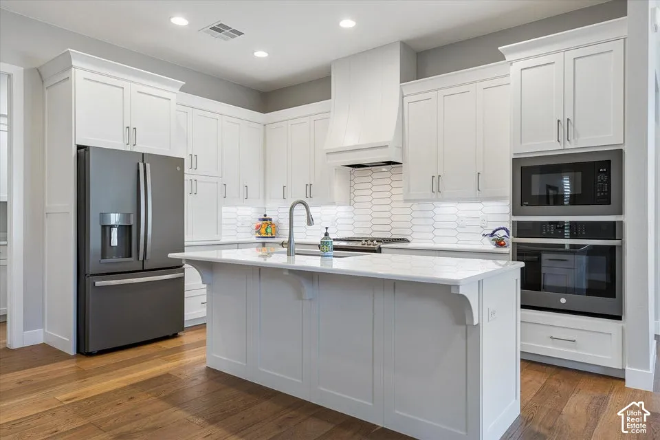White shaker-style cabinetry, stainless steel appliances, central island with sink, subway tile backsplash, and wood-finish flooring