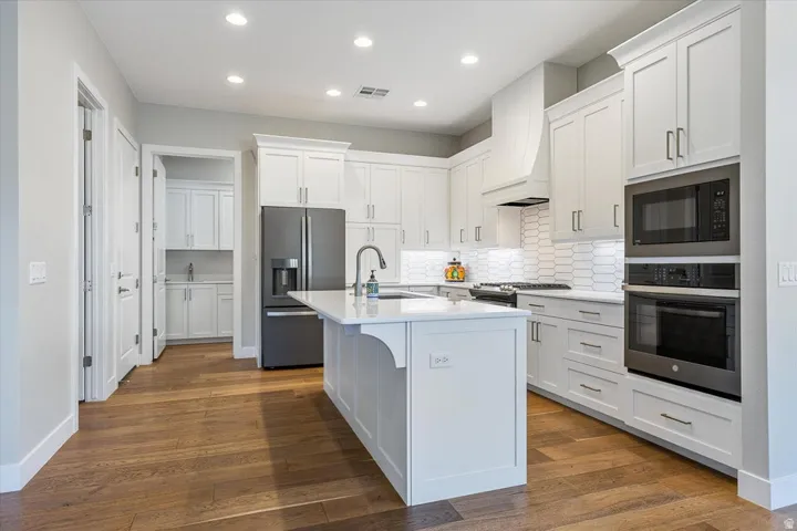 White shaker cabinetry, wood-finish flooring, recessed lighting, built-in stainless steel appliances, and a central island with a sink
