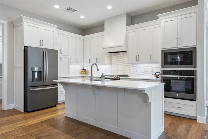 White shaker-style cabinetry, stainless steel appliances, central island with sink, subway tile backsplash, and wood-finish flooring