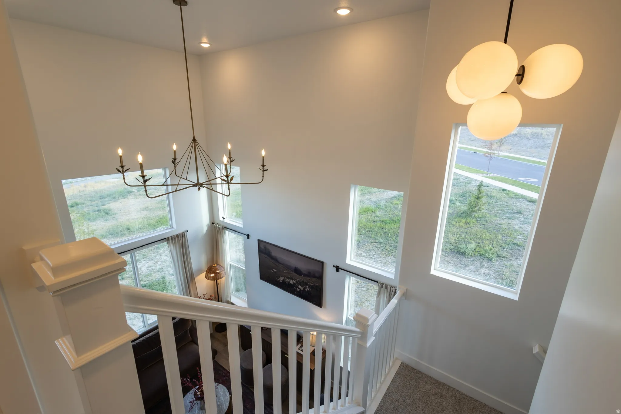 Soaring ceilings with recessed lighting, two distinct decorative chandeliers, multiple large windows, and a white wood-finish staircase with carpeted treads