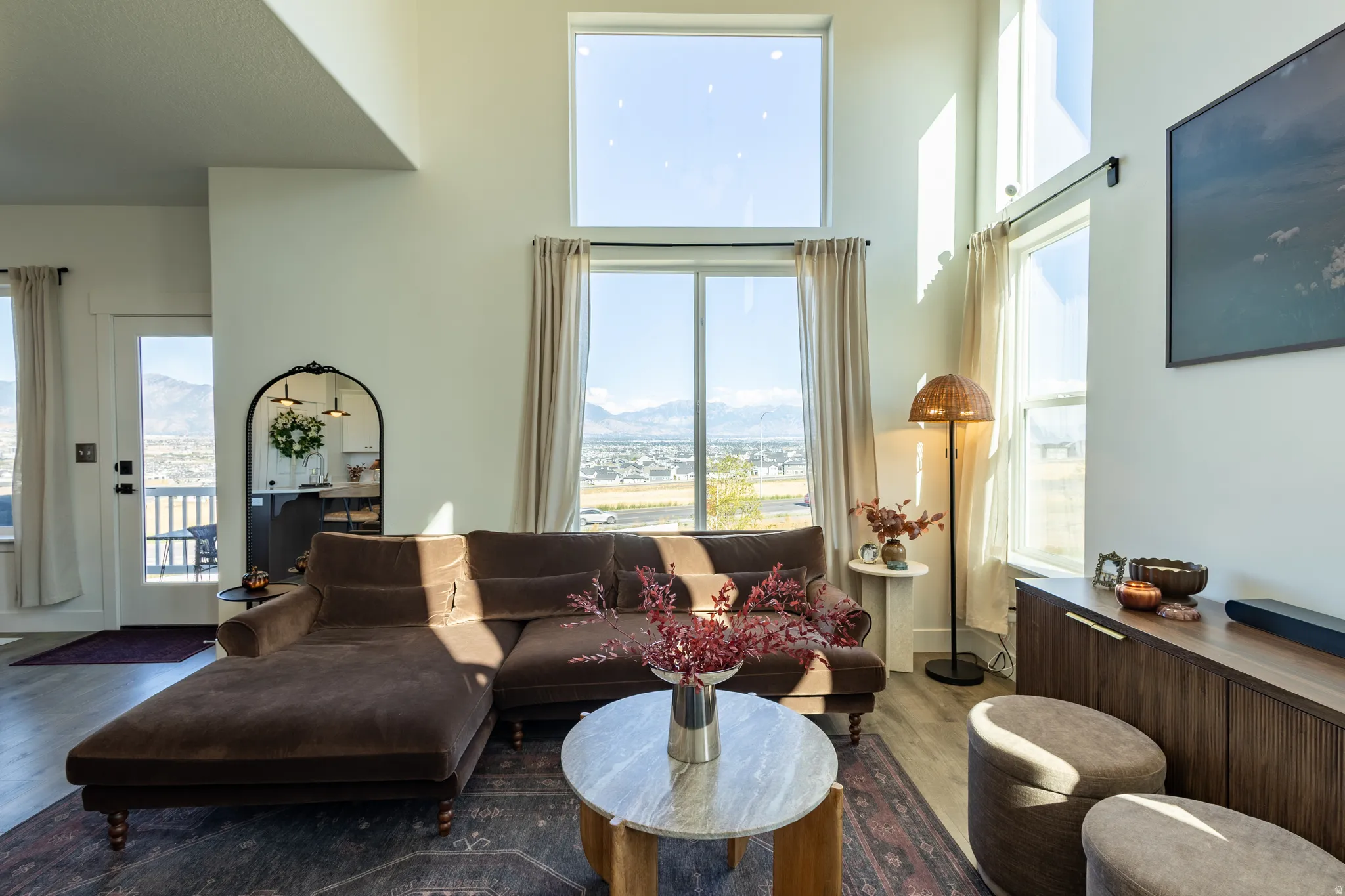 Sunlit living room showcasing floor-to-ceiling windows with expansive scenic vistas, rich charcoal upholstered sectional with chaise lounge, marble-top round cocktail table, wide-plank wood-finish flooring, and brass floor lamp with warm ambient lighting