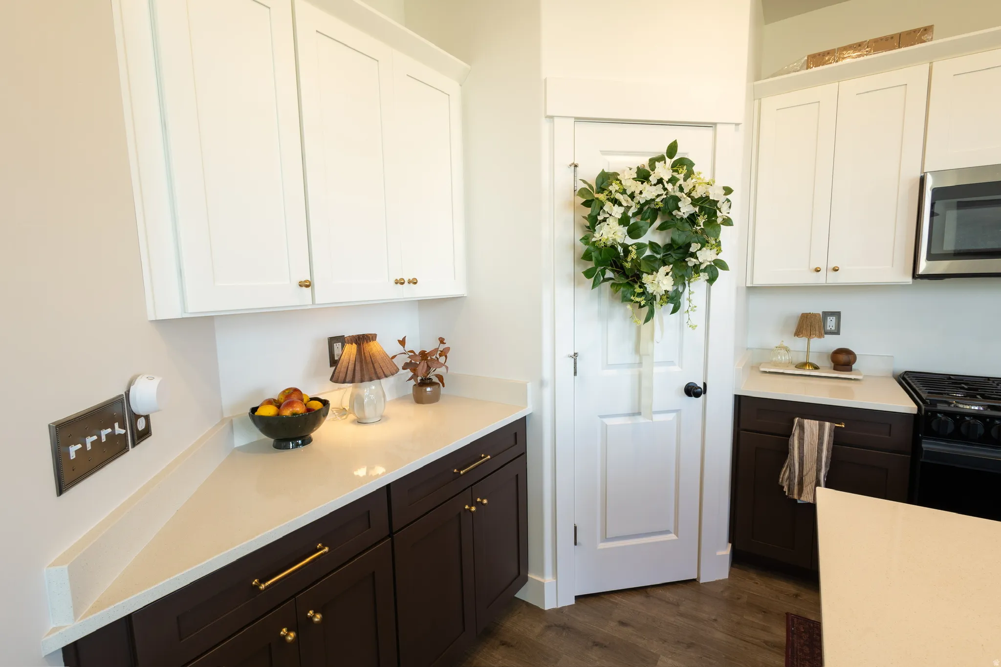 White upper cabinetry with gold-tone hardware, dark wood-finish lower cabinetry, light-toned countertops, wood-finish flooring, and a stainless steel microwave