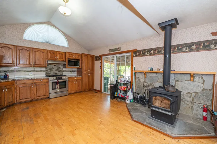 Vaulted ceiling featuring an arched window, wood-finish flooring, stainless steel appliances, and a wood-burning stove with a natural stone surround